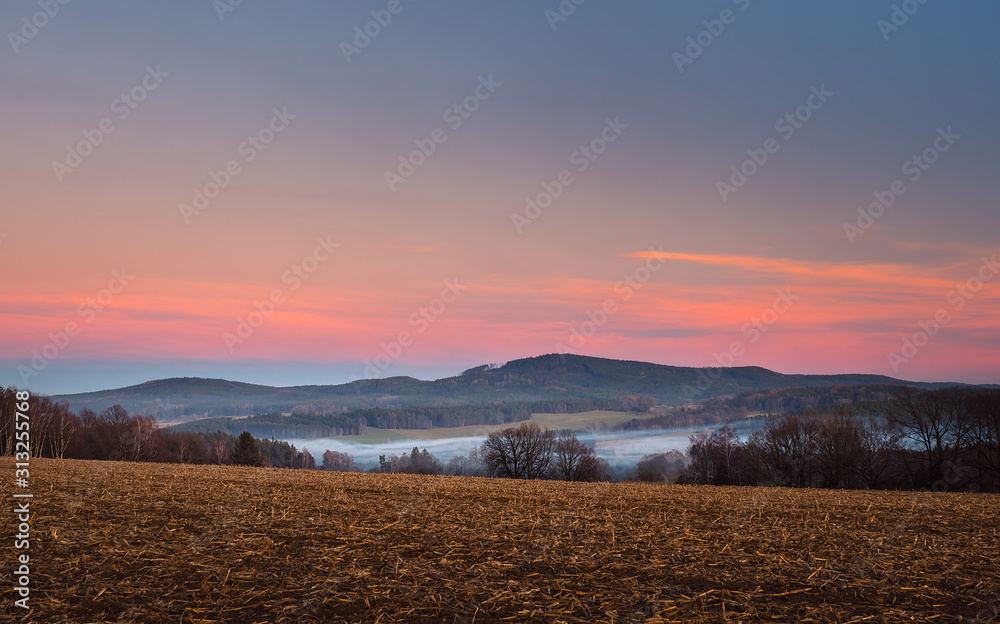 landscape at sunset, in the foreground field and trees, in the valley white fog, in the background forest and mountains, red and blue sky landscape at sunset, in the foreground field and trees, in the valley white fog, in the background forest and mountains, red and blue sky