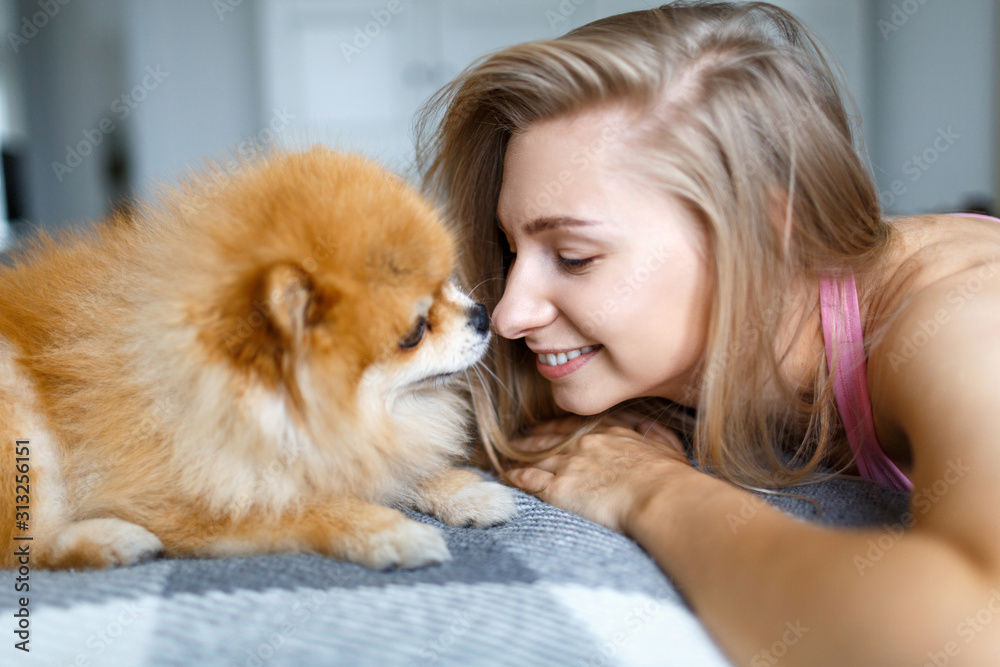 Happy blonde woman is lying on the bed with her dog of breed Spitz. Portrait of a woman and a dog.