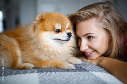 Happy blonde woman is lying on the bed with her dog of breed Spitz. Portrait of a woman and a dog. © Kanstantsinzzz