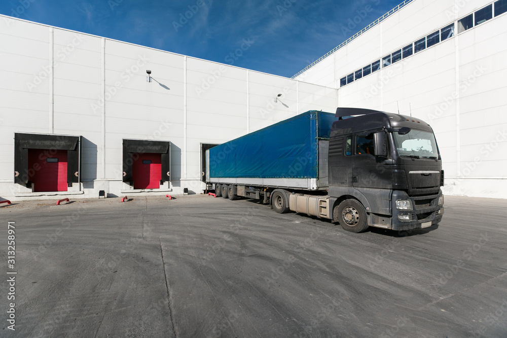 black truck with a blue trailer at unloading at the warehouse.Warehouse complex with an asphalt pad and truck with blue sky