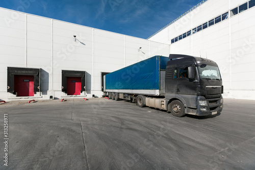 black truck with a blue trailer at unloading at the warehouse.Warehouse complex with an asphalt pad and truck with blue sky