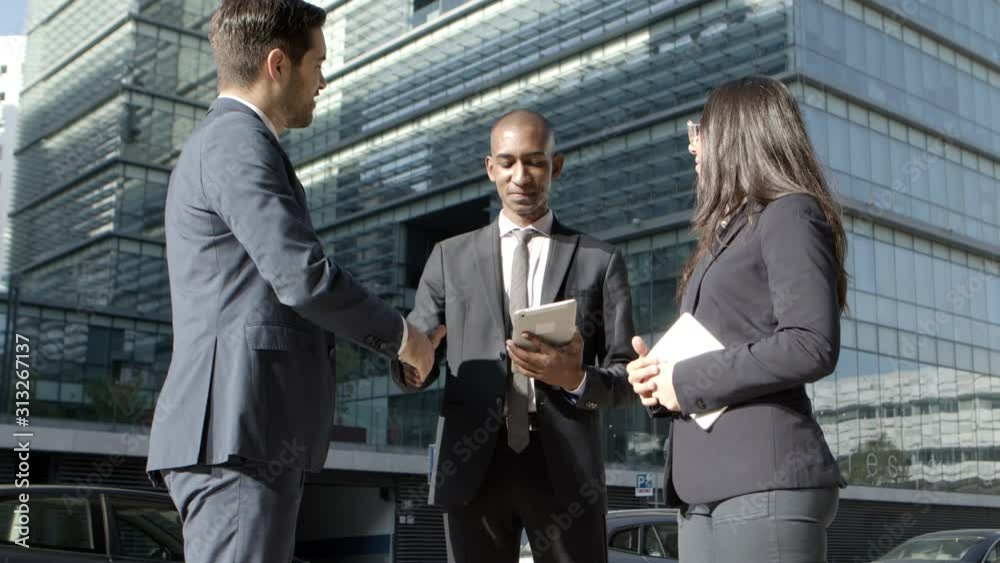 Coworkers with digital devices meeting on street. Multiethnic young businessmen and businesswoman with gadgets shaking hands and discussing work while meeting on street. Business cooperation concept