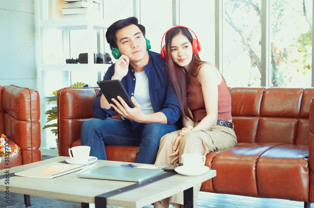 Asian handsome man and beautiful woman lovely couple choose music on red tablet device for listen on headphone setting on brown leather sofa in home with cute balloon decoration for winter party. Asian handsome man and beautiful woman lovely couple choose music on red tablet device for listen on headphone setting on brown leather sofa in home with cute balloon decoration for winter party.