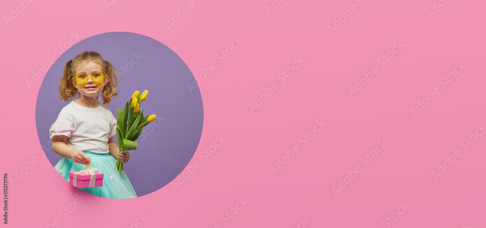 Obraz premium Adorable smiling child with spring flower bouquet and greating card in circle hole in the wall. Little toddler girl holding yellow tulips as gift for mother. 8 march or mother day concept