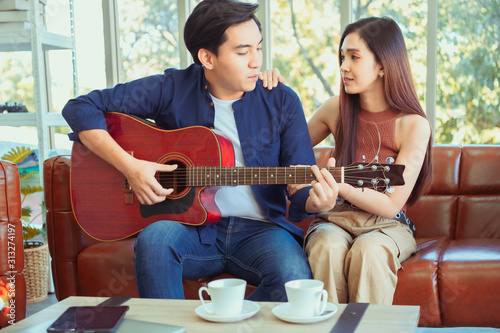 Young couple in love sitting on the sofa while young man playing guitar in living room. Happy couple in love. Stunning sensual portrait of young stylish fashion couple indoors.
