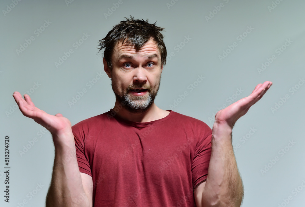 Portrait of a wild unkempt unshaven middle-aged man of 40 years in a burgundy t-shirt on a gray background. He stands right in front of the camera, talking, showing emotions. Waves his hands. Portrait of a wild unkempt unshaven middle-aged man of 40 years in a burgundy t-shirt on a gray background. He stands right in front of the camera, talking, showing emotions. Waves his hands.