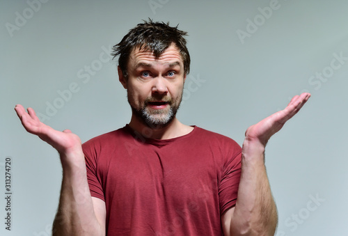 Portrait of a wild unkempt unshaven middle-aged man of 40 years in a burgundy t-shirt on a gray background. He stands right in front of the camera, talking, showing emotions. Waves his hands.