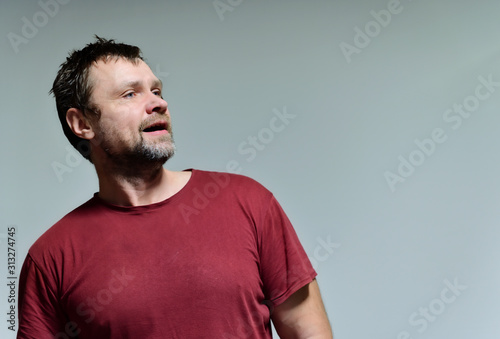 Portrait of a wild unkempt unshaven middle-aged man of 40 years in a burgundy t-shirt on a gray background. He stands right in front of the camera, talking, showing emotions. Waves his hands.