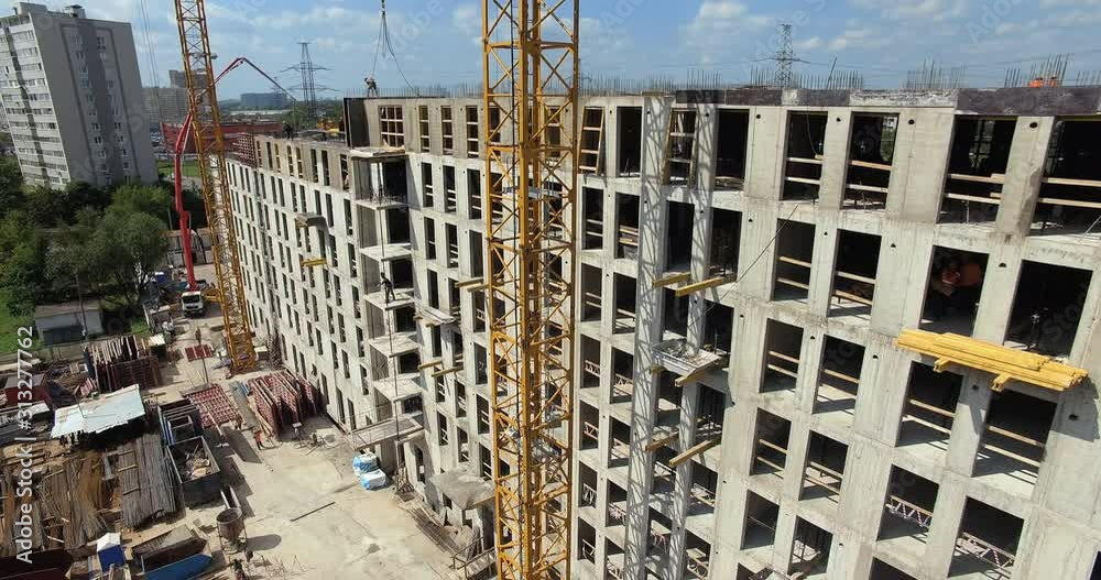 A vertical inspection of construction site of a multi storey building on a sunny summer day. Concrete walls with empty window holes, yellow construction cranes near the facade and busy builders on A vertical inspection of construction site of a multi storey building on a sunny summer day. Concrete walls with empty window holes, yellow construction cranes near the facade and busy builders on