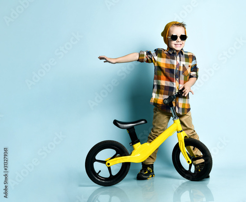 Smiling boy in bright stylish casual clothing, sneakers, cap and round sunglasses standing and holding his yellow run bike present