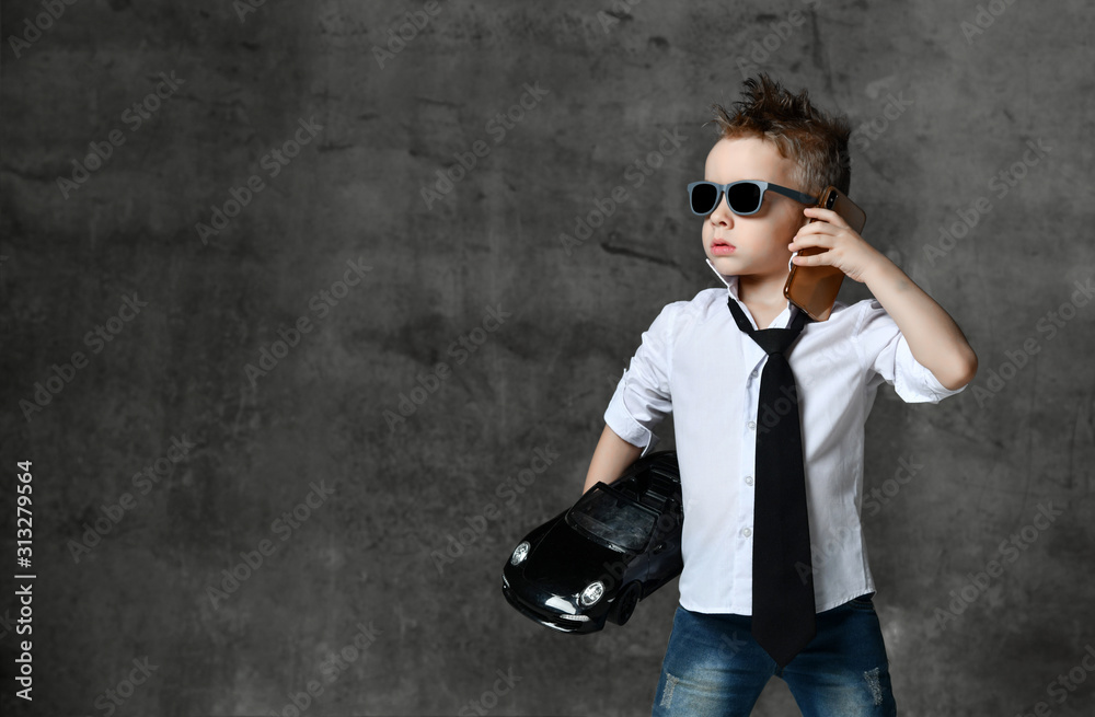 Serious boy in stylish white shirt, jeans, tie and sunglasses standing, holding black toy car present in hand and calling on phone