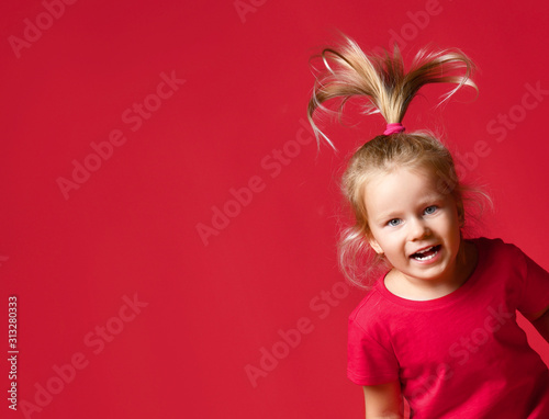 Portrait of jumping small happy excited blond girl in stylish casual clothing with hair raised up over red wall background