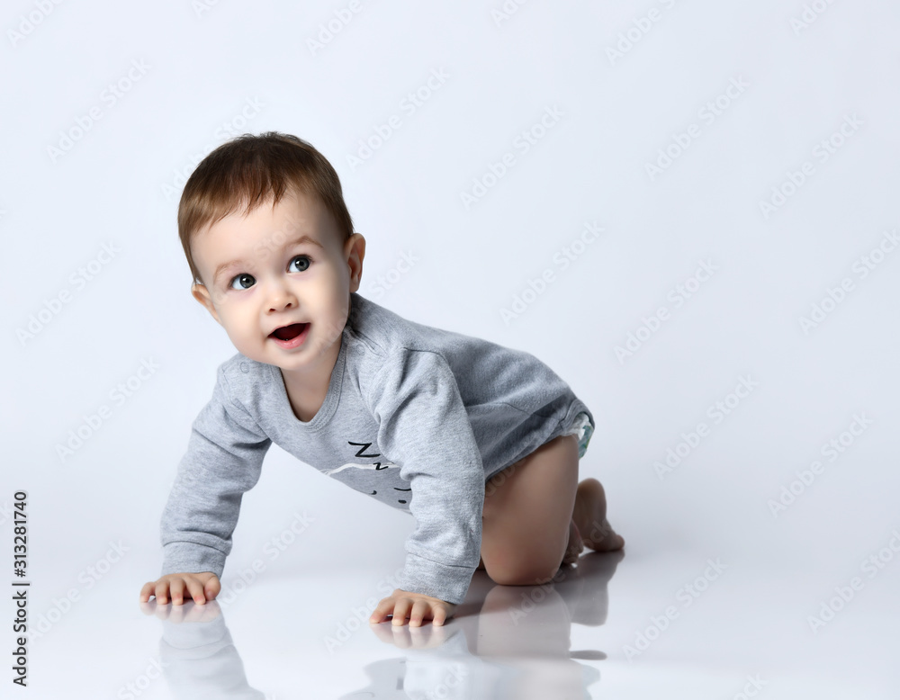 Little baby boy toddler in grey casual jumpsuit and barefoot crawling on floor, smiling and looking up over white wall background Little baby boy toddler in grey casual jumpsuit and barefoot crawling on floor, smiling and looking up over white wall background
