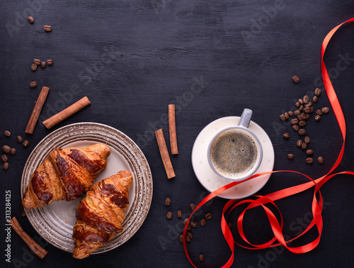 Croissants on a ceramic plate, a cup of coffee, red ribbon, cinnamon sticks and coffee beans on a black wooden table. Top view with space for text. Background for restaurant, bakery, cafe.