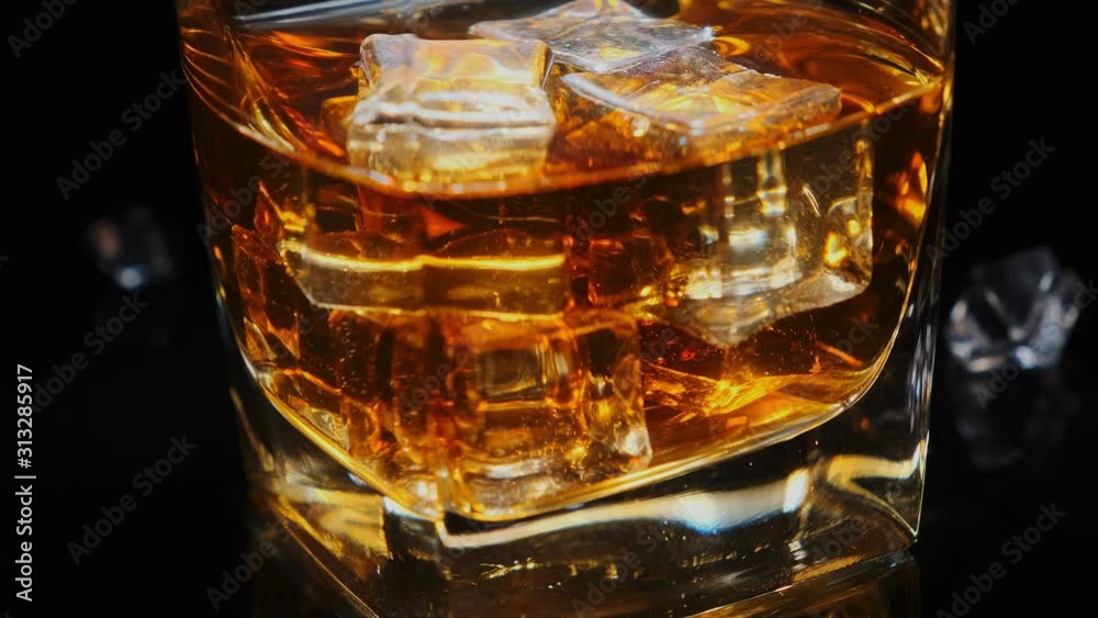 Golden whiskey in faceted glass with ice cubes rotating on black background macro. Alcoholic drinks, cocktail, alcohol. Golden whiskey in faceted glass with ice cubes rotating on black background macro. Alcoholic drinks, cocktail, alcohol.