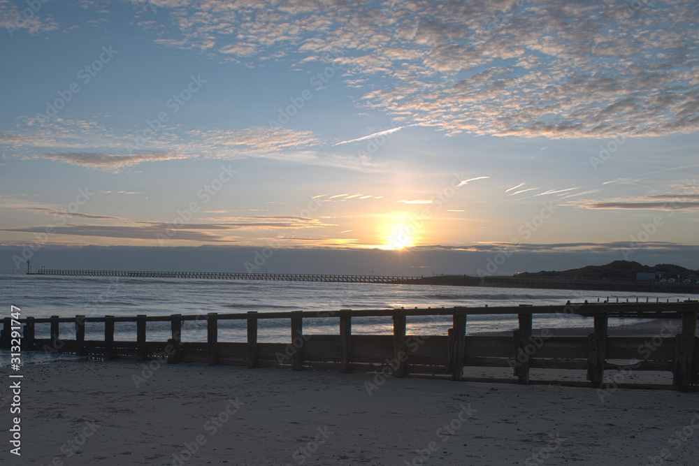 Obraz premium Beautiful Sunset on Littlehampton seafront as the sun dips behind cloud on the horizon, reflecting vibrant colours onto the clouds.