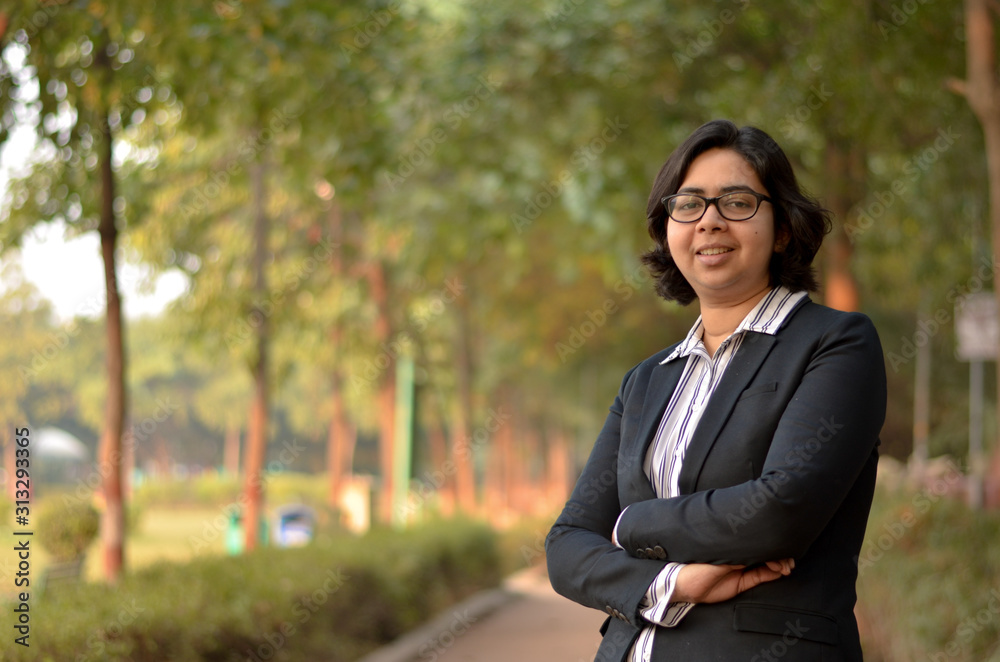 Closeup portrait of a confident young Indian Corporate professional woman  with short hair and spectacles, crossed folded hands in an outdoor setting  wearing a black business / formal suit Stock Photo |