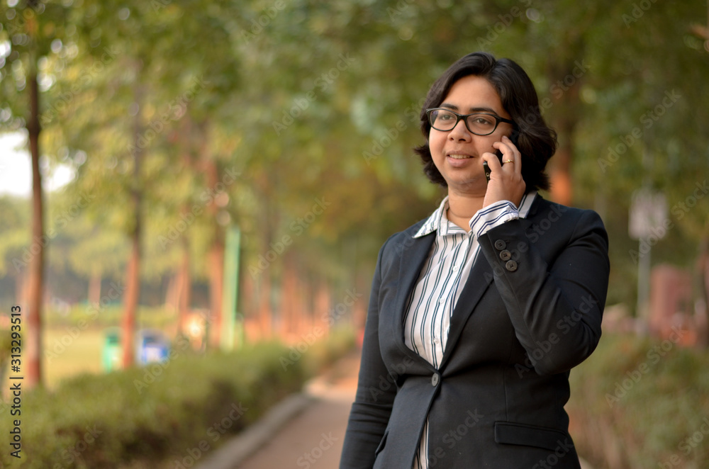 Portrait of a confident looking young Indian woman with short hair and spectacles speaking on a mobile phone and holding a laptop in outdoor setting wearing a formal striped shirt in the park Portrait of a confident looking young Indian woman with short hair and spectacles speaking on a mobile phone and holding a laptop in outdoor setting wearing a formal striped shirt in the park