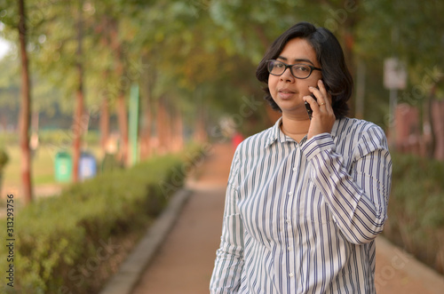 Portrait of a confident looking young Indian woman with short hair and spectacles speaking on a mobile phone and holding a laptop in outdoor setting wearing a formal striped shirt in the park