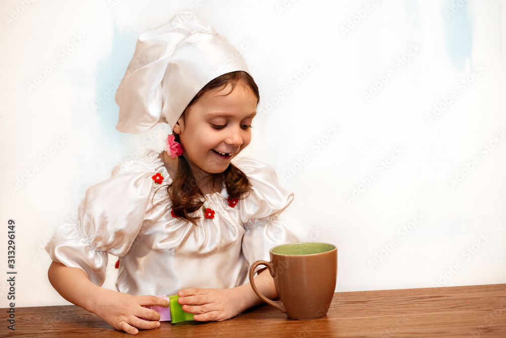 The girl in the chefs hat is at the kitchen table and looks in the cup The girl in the chefs hat is at the kitchen table and looks in the cup
