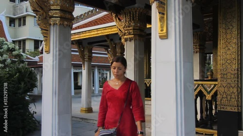 A young woman wearing a red dress is walking around a Buddhist temple in Bangkok, Thailand. The camera dollies to the left side. (ID: 313297518)