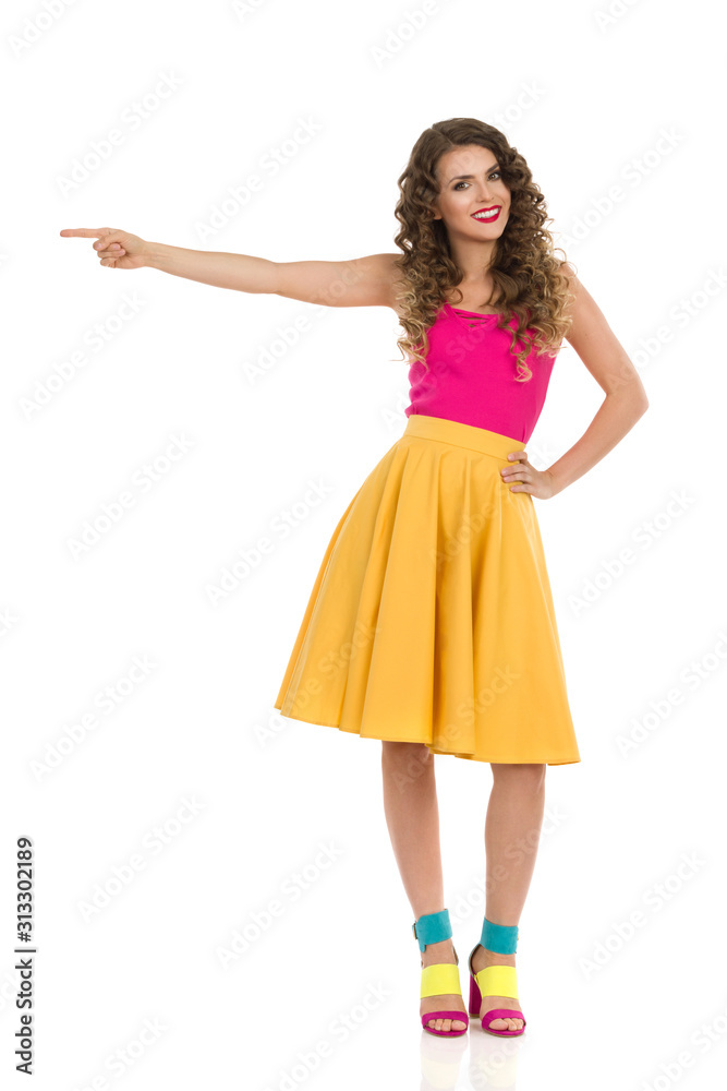 Cute Young Woman In Colorful High Heels, Vibrant Skirt And Pink Top Is Pointing At The Side.