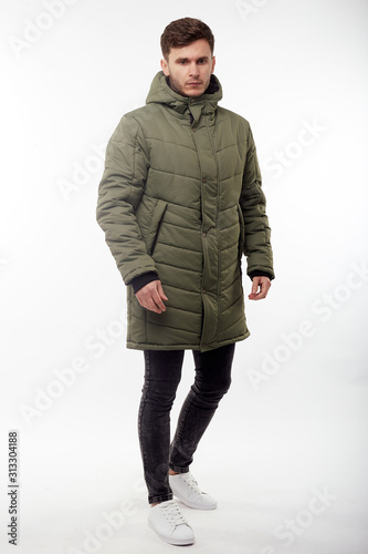 Young european man in white sweater and black pants, olive down jacket posing on white background. Isolated.