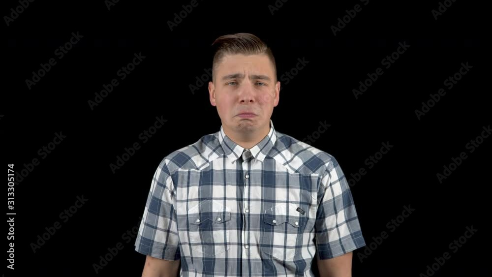 A young man takes off his mask and shows an emotion of sadness on his face.. Sad man in a shirt. A man hides his face behind a white mask on a black background. A young man takes off his mask and shows an emotion of sadness on his face.. Sad man in a shirt. A man hides his face behind a white mask on a black background.