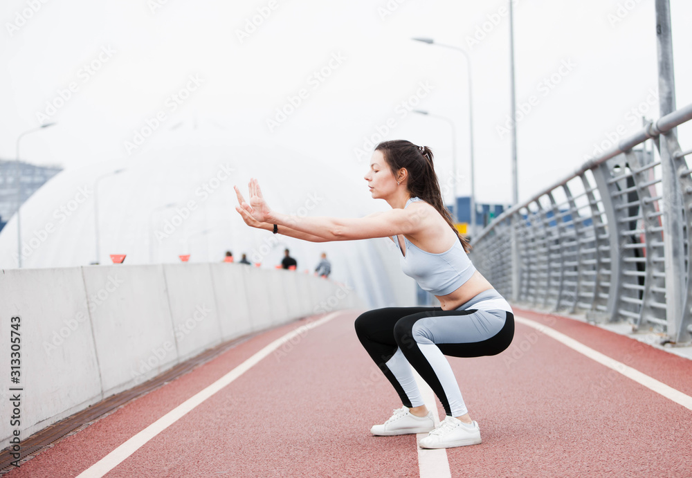 Young sports woman in fitness suit crouches, lunges, plays sports against the backdrop of urban cityscape and white sky. Young sports woman in fitness suit crouches, lunges, plays sports against the backdrop of urban cityscape and white sky.