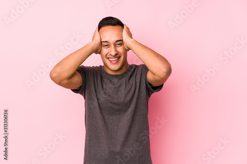 Yooung latin man posing in a pink backgroundlaughs joyfully keeping hands on head. Happiness concept.