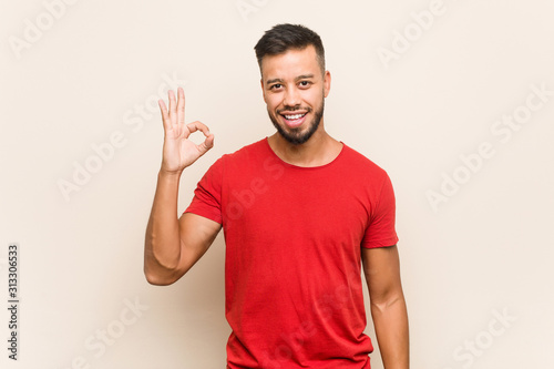Young south-asian man winks an eye and holds an okay gesture with hand.