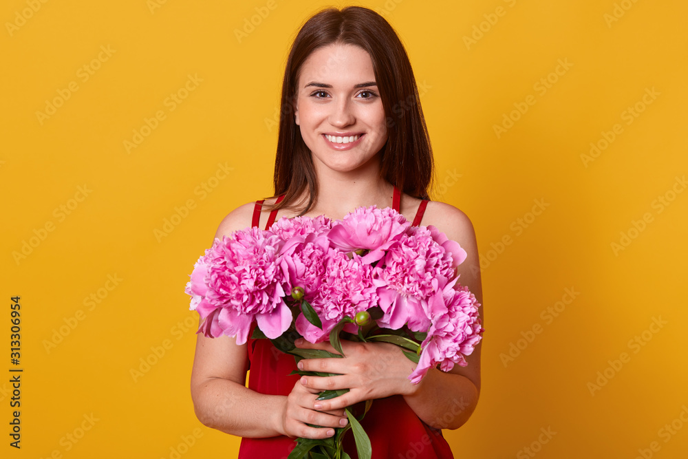 Close up portrait of gorgeous brunette woman with perfect peony flower, posing isolated over yellow background, female looking smiling directly at camera, embracing her bouquet of pink peonies.