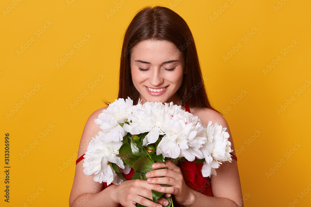 Indoor shot of young cute woman smelling white flowers,looking down at her bouquet, holding gentle peonies in hands, attractive smiling lady posing with flowers against yellow studio background. Indoor shot of young cute woman smelling white flowers,looking down at her bouquet, holding gentle peonies in hands, attractive smiling lady posing with flowers against yellow studio background.