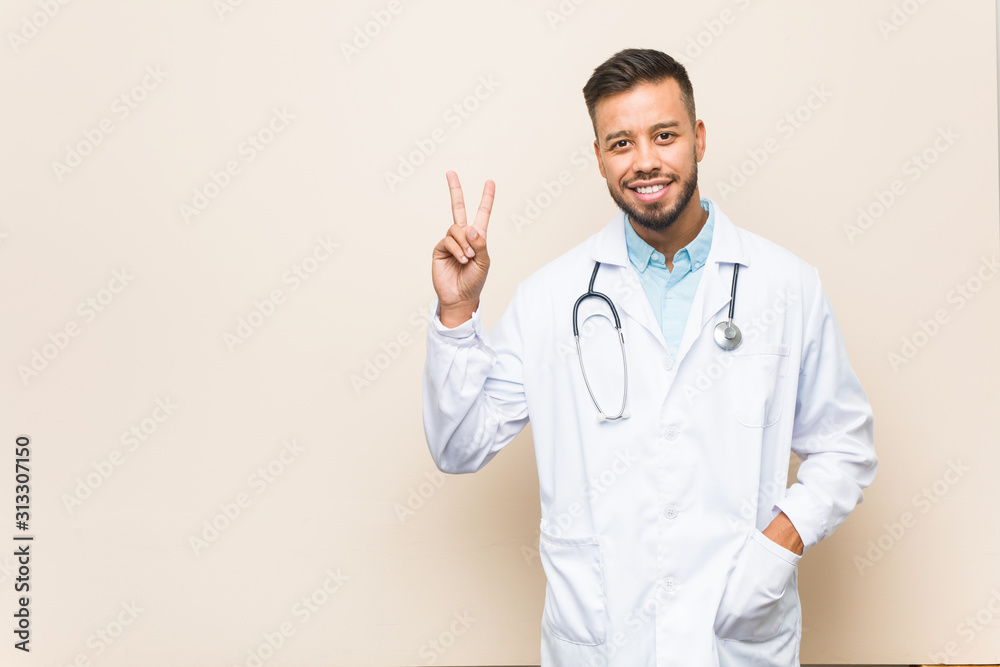 Young south-asian doctor man joyful and carefree showing a peace symbol with fingers.