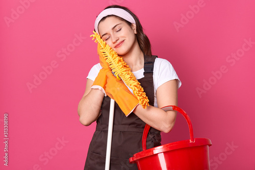Beautiful housewife being tired to clean up room, standing with closed eyes, falling asleep, being pleased but tired, posing isolated over pik studio background. Housekeeping and cleaning concept. © sementsova321