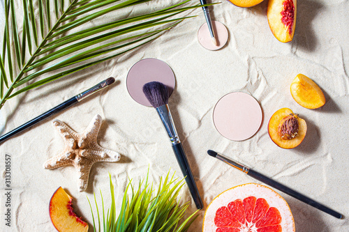 Dry cosmetic decorative product in round refile lies on the sand, surrounded by bright fruits, corals, and palm leaves. photo