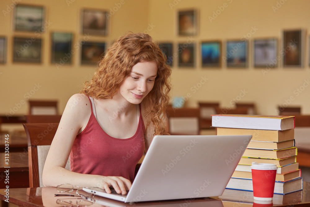 Young pretty female student with laptop and books working in high school library, sitting at table, takeaway cooffe near her books, woman with wavy hair wearing maroon t shirt. Education concept. Young pretty female student with laptop and books working in high school library, sitting at table, takeaway cooffe near her books, woman with wavy hair wearing maroon t shirt. Education concept.