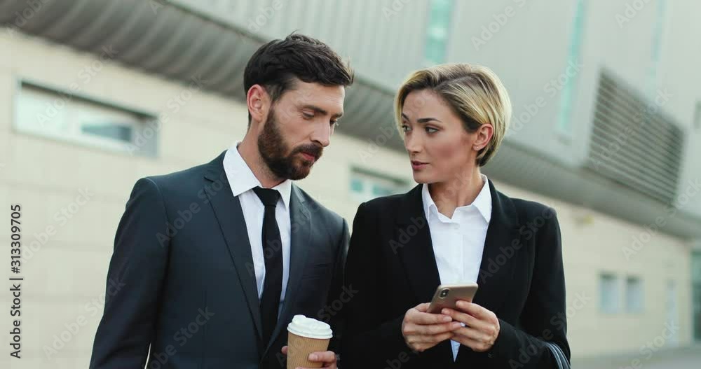 Caucasian handsome businessman drinking coffee to-go and watching how his female colleague tapping and scrolling on the smartphone while shwing something to him. Caucasian handsome businessman drinking coffee to-go and watching how his female colleague tapping and scrolling on the smartphone while shwing something to him.