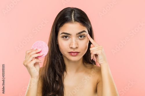 Young indian woman holding a facial skin care disc pointing his temple with finger, thinking, focused on a task.