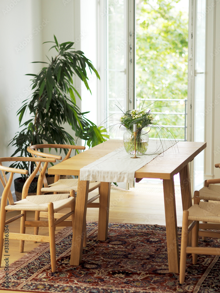 Fresh flowers in glass vase standing on a wooden table in white dining room interior with oriental carpet. Interior design in minimal style.