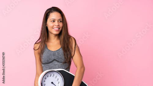 Young sport Brazilian girl over isolated pink background with weighing machine