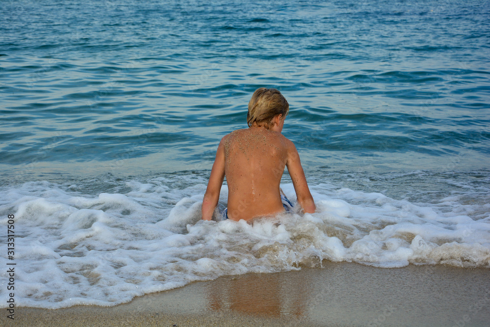 Fototapeta premium A blond boy from behind, sitting in a wave on a sandy beach