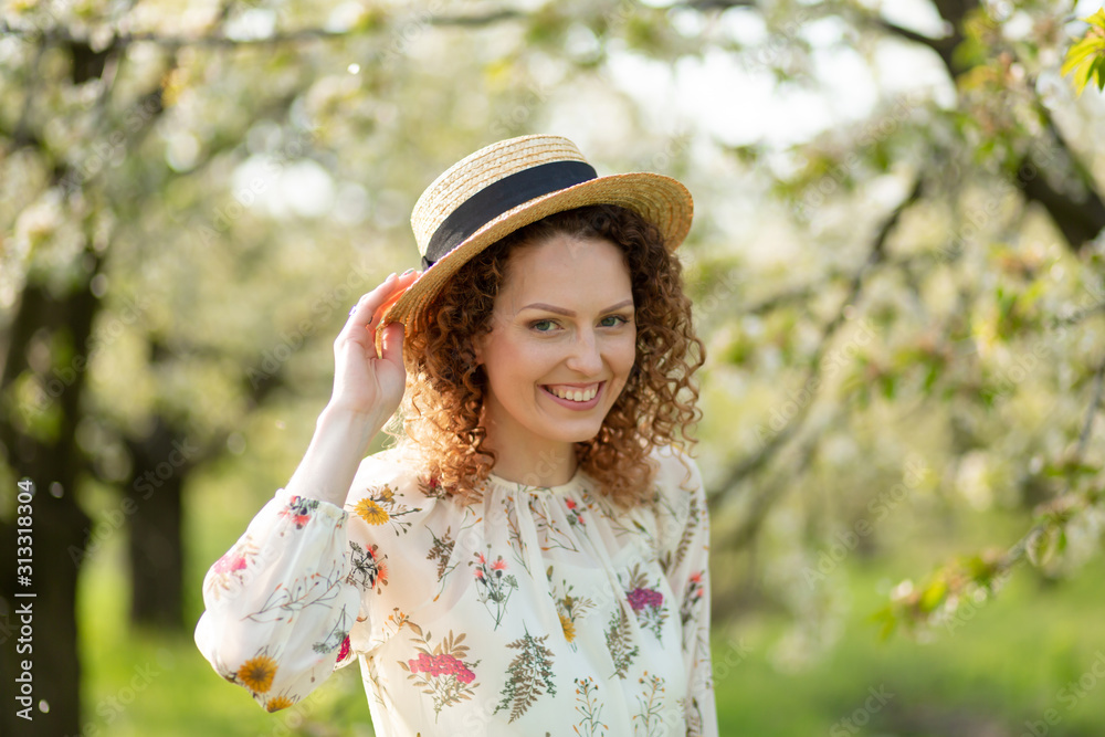 Portrait young attractive woman with curly hair in stylish wicker hat enjoys blooming green garden in spring day