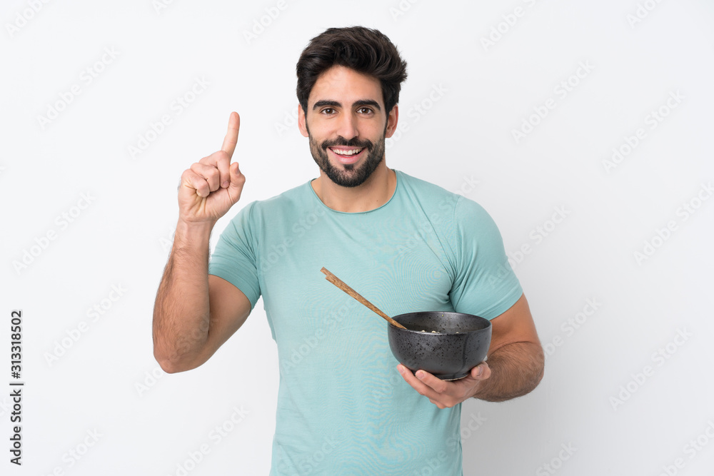 Young handsome man with beard over isolated white background pointing up a great idea while holding a bowl of noodles with chopsticks Young handsome man with beard over isolated white background pointing up a great idea while holding a bowl of noodles with chopsticks