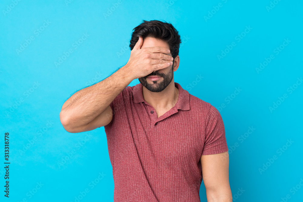 Young handsome man with beard over isolated blue background covering eyes by hands. Do not want to see something Young handsome man with beard over isolated blue background covering eyes by hands. Do not want to see something