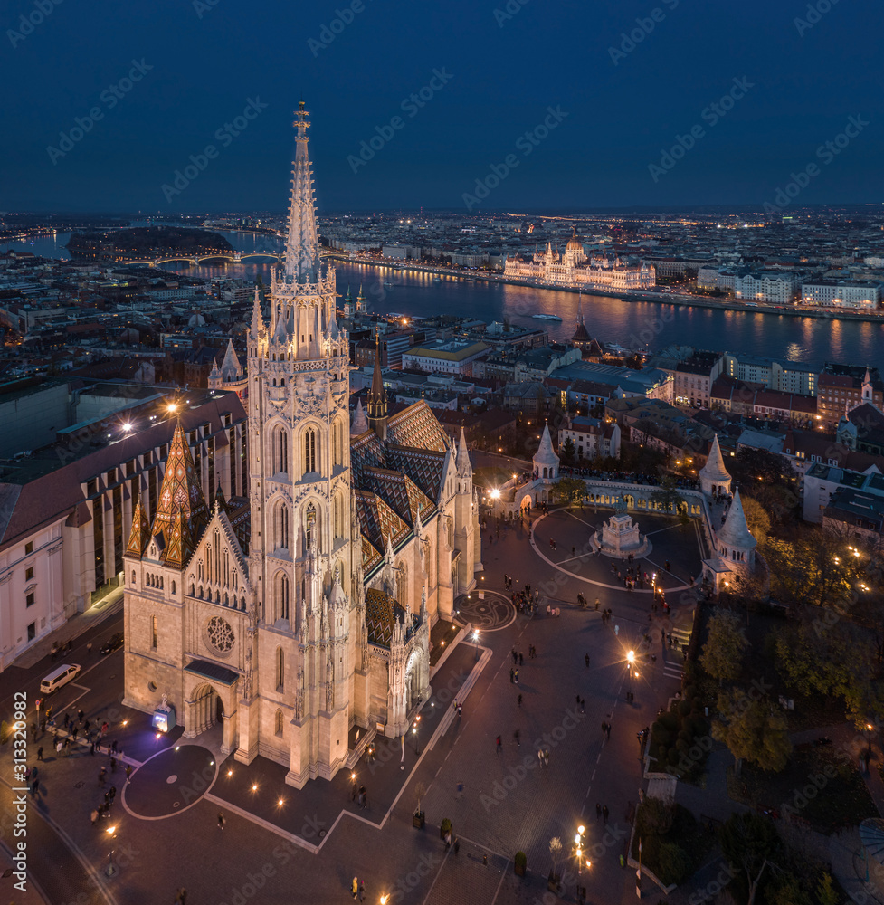 Naklejka premium Budapest, Hungary - Aerial drone view of the beautiful illuminated Matthias Church at blue hour with Fisherman's Bastion (Halaszbastya) and Parliament of Hungary at background