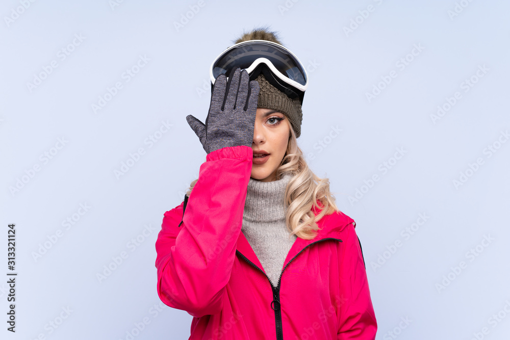 Skier teenager girl with snowboarding glasses over isolated blue background covering a eye by hand