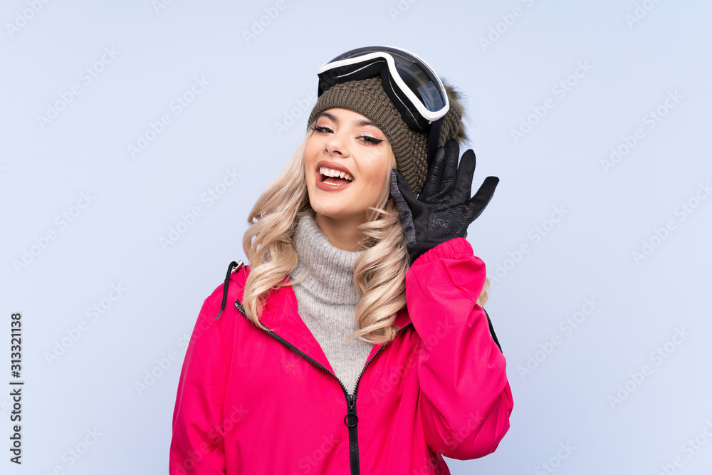 Skier teenager girl with snowboarding glasses over isolated blue background listening something