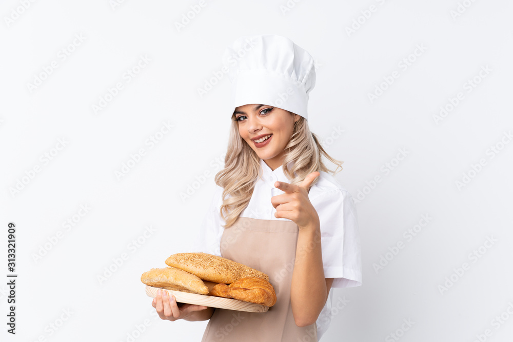 Teenager girl in chef uniform. Female baker holding a table with several breads over isolated white background points finger at you Teenager girl in chef uniform. Female baker holding a table with several breads over isolated white background points finger at you