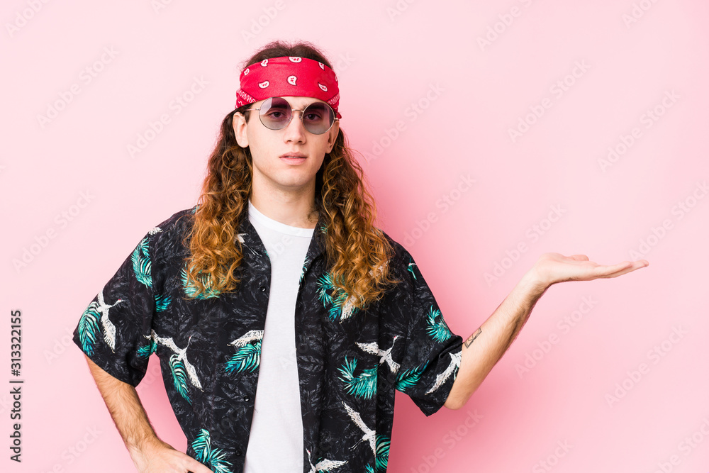 Young long hair hippie man posing in a pink background showing a copy space on a palm and holding another hand on waist.